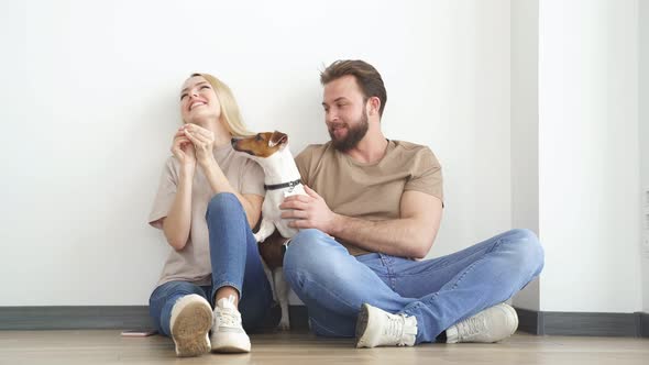 Young Happy Couple is Moving Into New House Sitting Down on Floor with Little Puppy