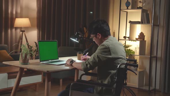 Asian Man Sitting In A Wheelchair While Working On Green Screen Laptop Computer And Note In Book alt