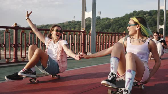 Portrait of Two Teenage Girls Riding on City Bridge Sitting on Skateboards and Holding Hands Youth alt