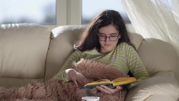 Funny Girl in Glasses Lying on the Sofa at Home Reading a Book and Smiling