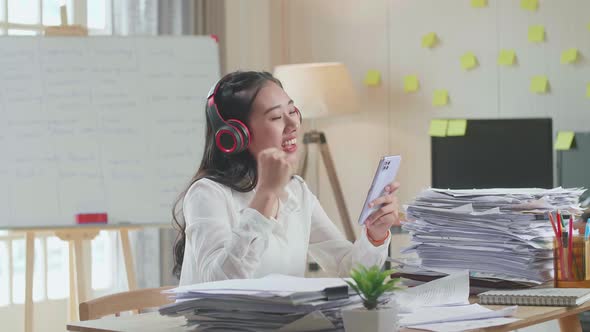 Asian Woman With Headphones Listening To Music On Smartphone After Working With Documents alt