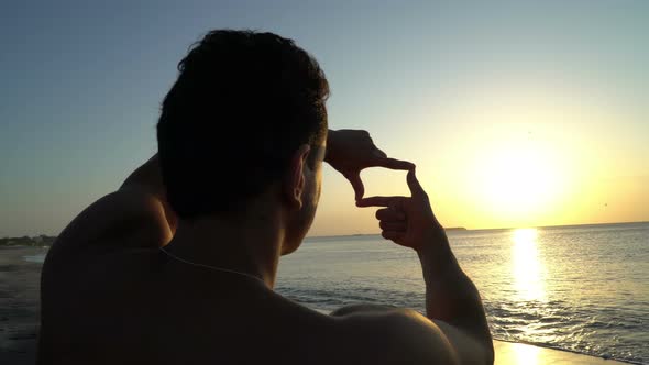 Young Man with Curly Hair Making Photo Frame with His Hands at Amazing Sunset on the Beach