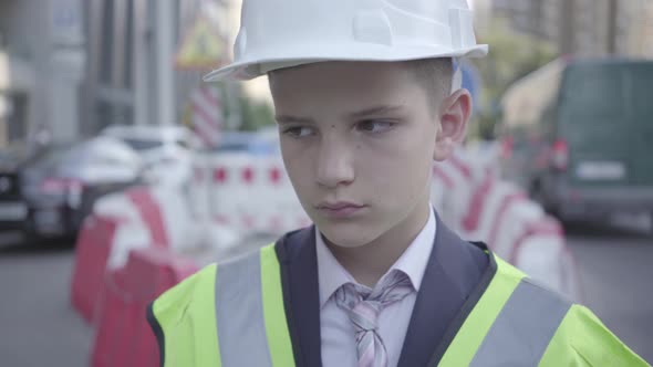 Portrait of Sad Tired Little Boy in Constructor Helmet on His Head, and Uniform Looking Away alt