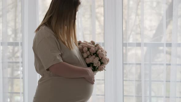 Pregnant Woman Stroking Her Big Belly and Holding Bouquet of Rose Flowers on the Background of