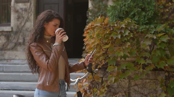 Pretty young woman with mobile phone drink coffee to go by the bicycle on autumn day