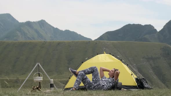 Girl Tourist with Gray Clothes Relaxes Lying in Front of the Tent on a Background of Mountains