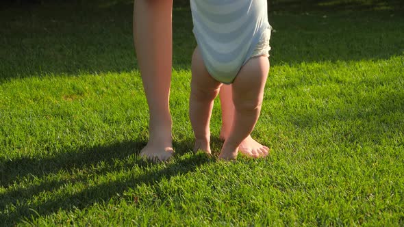 Closeup of Barefoot Baby Boy and Mother Standing on Fresh Green Grass at Park alt