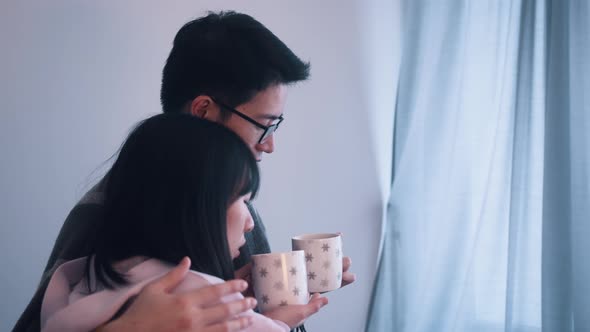 Young Couple Hugging Near the Window with Hot Tea on Stormy Day alt