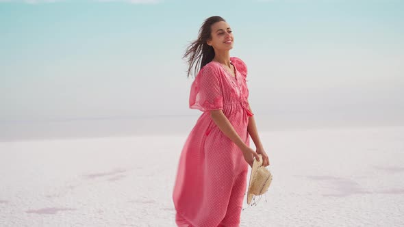 Woman in Blowing Airness Pink Dress and Hat Standing on Salty Beach on Pink Lake alt
