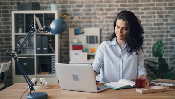 Young Woman Employee Using Laptop and Writing in Notebook at Desk in Office alt