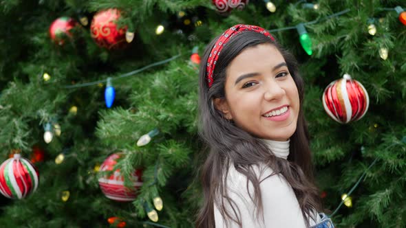 A gorgeous hispanic woman smiling and looking merry celebrating Christmas with a tree and festive ho alt