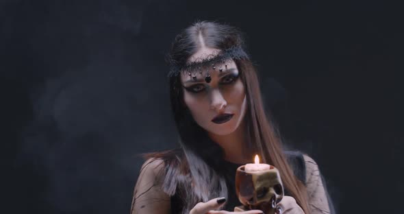 Mystical Female in Black Attire Holds a Skull with a Burning Candle and Looking at the Camera, the alt