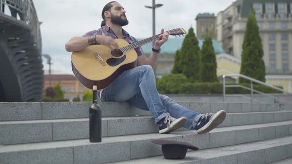 Wide Shot Portrait of Enthusiastic Musician Playing Guitar on City Stairs with Wine Bottle and Hat alt
