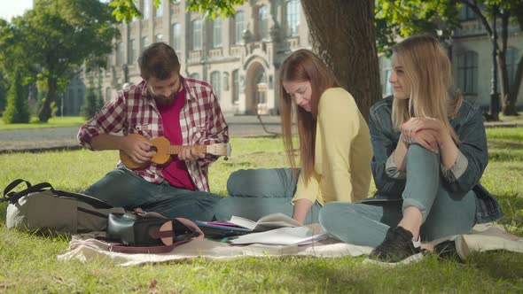 Positive Caucasian Man Playing Ukulele for Beautiful Girls on Sunny Green Meadow. Portrait of Young alt