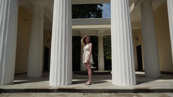 A Woman in a White Dress Poses Standing Between Two Tall White Columns alt