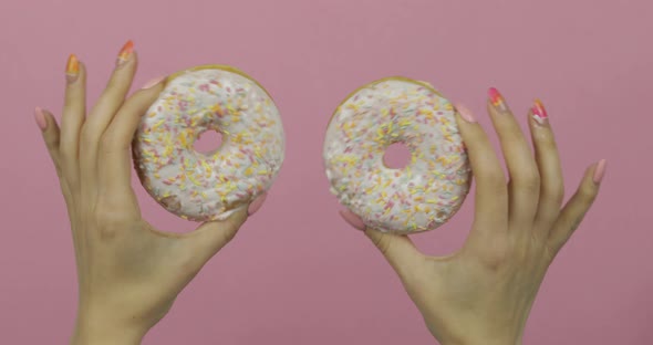 Womens Hands Holding Two White, Delicious, Sprinkled Donuts on Pink Background. alt