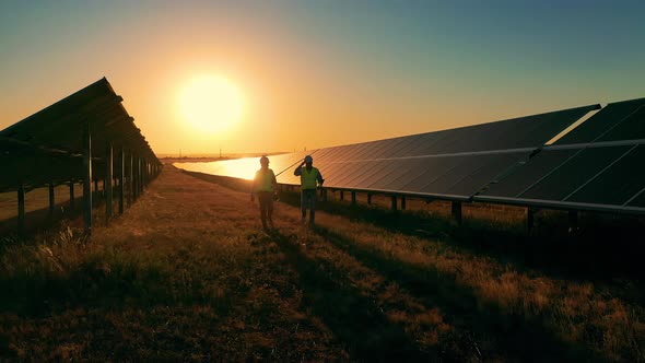 Two Electricians Passing By Long Rows of Solar Panels at Sundown alt