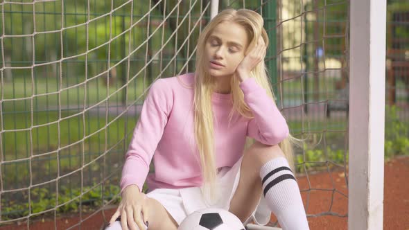 Charming Young Blond Woman Sitting with Football Ball on Outdoor Sports Ground. Camera Approaches To alt