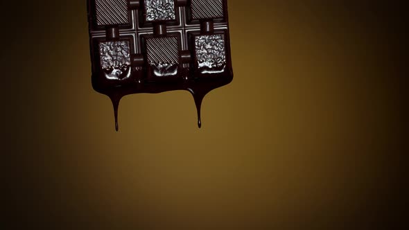 Slow Motion of Chocolate Bar with Melted Dark Chocolate Dripping Flowing Over Brown Background alt
