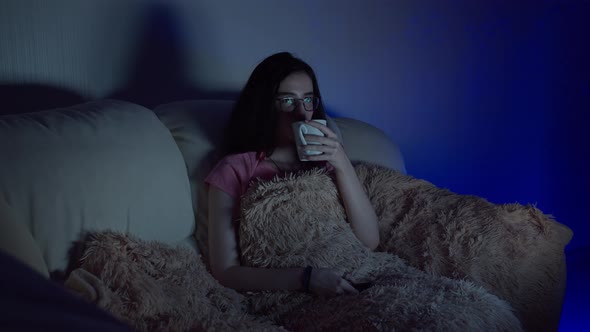 Girl with a Cup of Tea Sits on the Sofa in the Evening and Watches Tv Glare on Glasses