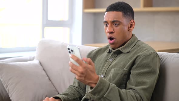 Worried Multiracial Millennial Guy Holding Smartphone Sitting on the Sofa