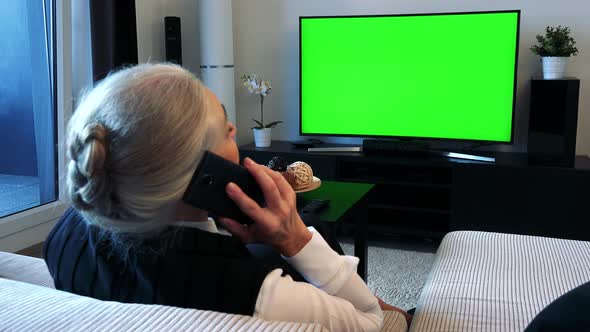 Old Caucasian Woman Watches Television in Living Room and Phone - Green Screen alt