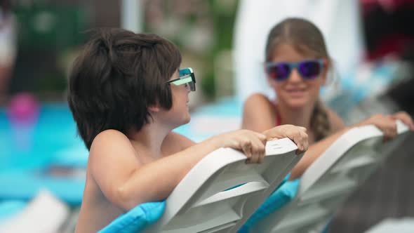 Cute Caucasian Boy in Sunglasses Posing on Sunbed with Blurred Sister Smiling at Background alt