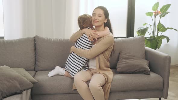 A Young Mother Gently Hugs Her Little Daughter Sitting on the Couch at Home alt