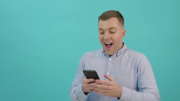 A Man in a Blue Shirt is Corresponding on a Smartphone and Expresses Great Joy and Surprise alt
