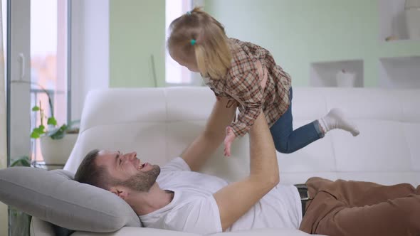 Side View of Cheerful Young Father Having Fun Holding Baby Girl Up Imitating Airplane As Joyful alt