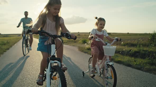 Caucasian family of two elementary age girls on foreground and parents in the background riding bike alt