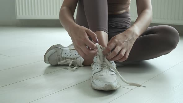 Young Girl in Sportswear Sits on Floor on Knees Untying Shoelaces on Sneakers alt