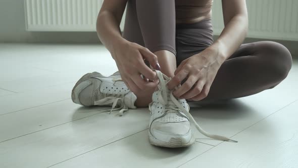 Young Girl in Sportswear Sits on Floor on Knees Untying Shoelaces on Sneakers