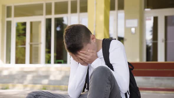 A Caucasian Teenage Boy Cries with His Face in His Hands As He Sits in Front of School  Closeup