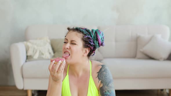Fat Woman in Sportswear Sitting on Mat Enjoying Eating Her Doughnut at Home