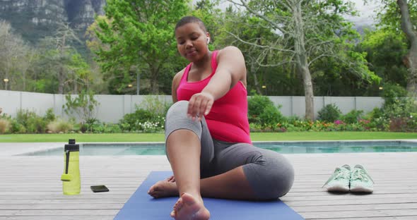 Happy african american plus size woman practicing yoga, stretching next to swimming pool in garden alt