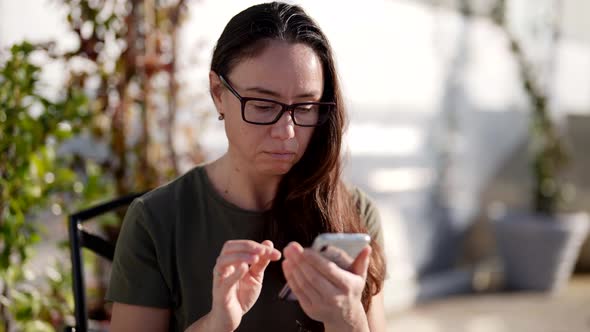 A Focused Woman with Glasses Writes a Message on Her Brand New Smartphone While Sitting at a Table alt