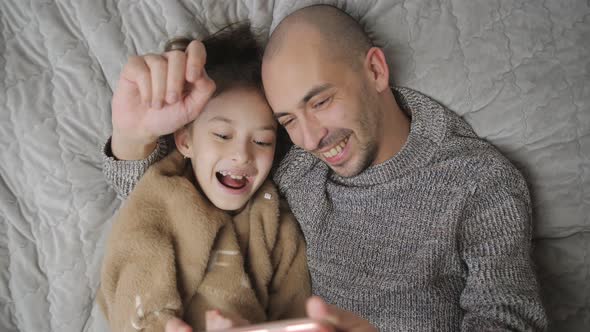 Happy Spending Time with a Father and Daughter at Home on the Bed Looking at a Smartphone alt
