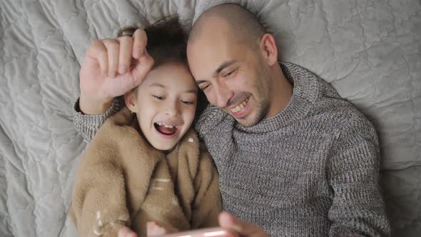 Happy Spending Time with a Father and Daughter at Home on the Bed Looking at a Smartphone