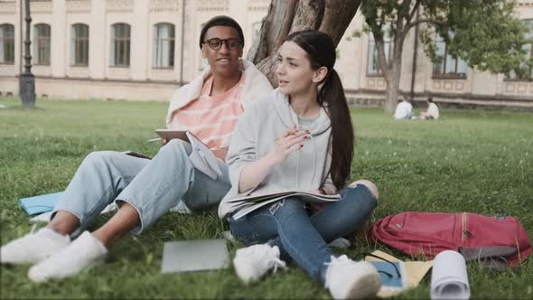 Couple of International Students Working on Homework Together in the Campus Sitting on the Lawn Near alt