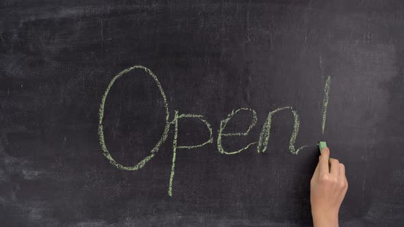 A woman's hand writes the word OPEN on a chalkboard with green chalk alt