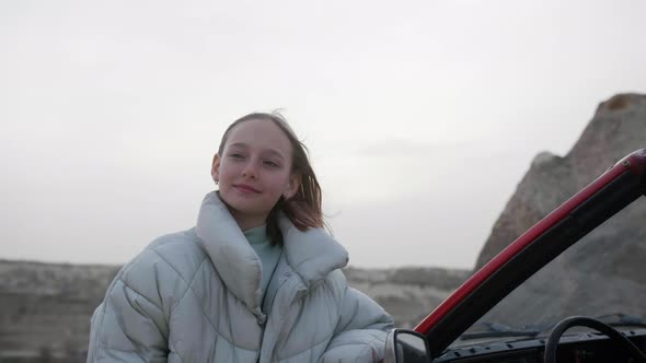 Attractive Teen Girl in Green Jacket Stand Near Red Loan Cabrio Car and Enjoy Landscape Desert Stone
