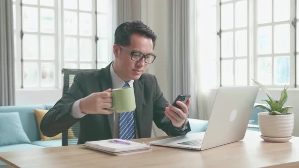 Asian Businessman Using Mobile Phone And Drinking Coffee While Using Computer Working At Home alt