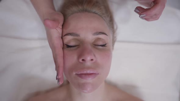 Headshot of Relaxed Beautiful Caucasian Woman Closing Eyes As Female Hands Massaging Face in Slow alt