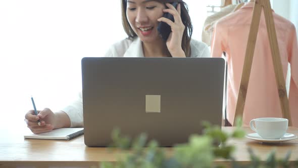 young smiling asian woman working on laptop while sitting in a living room at home using phone. alt