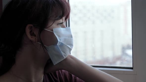 Woman With Mask On Face Looks Out Window At Street During Coronavirus Period alt