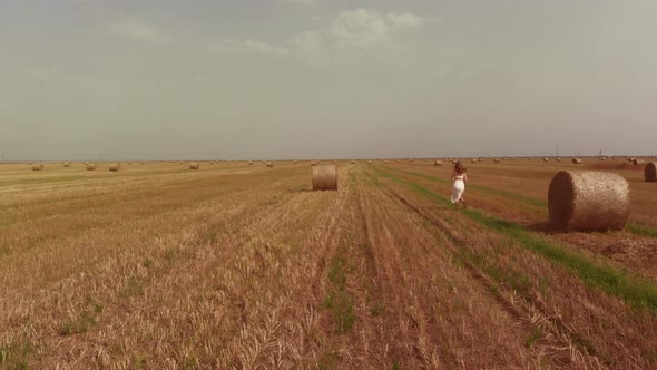 Woman in White Skirt Runs Along Field with Rolled Straw