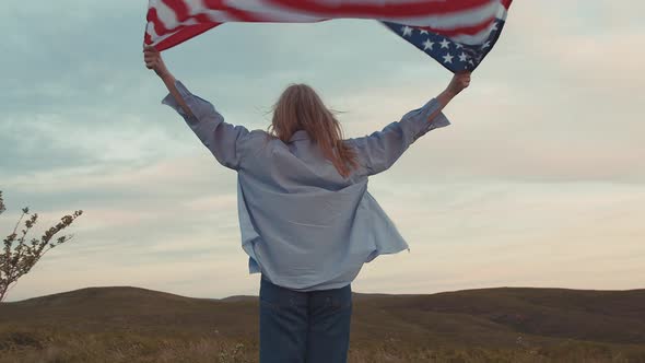 Blonde Woman Carries the American Flag with Hands Up and Walks Across the Field alt