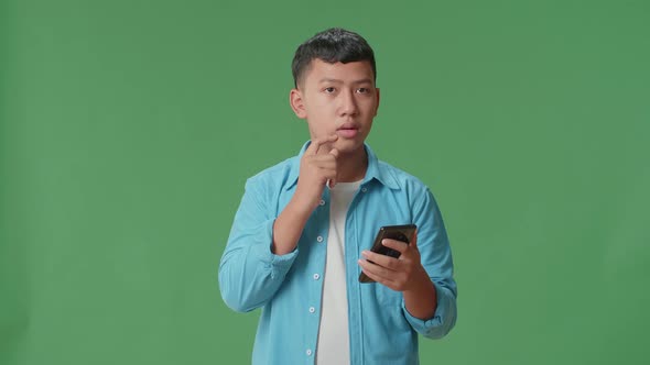 Young Asian Boy Use Mobile Phone And Thinking About Something While Standing On Green Screen alt