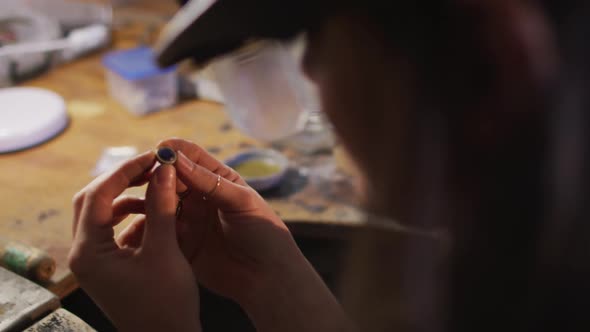 Close up of hands of caucasian female jeweller wearing glasses, checking jewelry at workshop alt